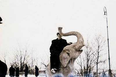 Black-and-white photo of a person perched atop a large, stylized elephant statue in an urban park. The statue’s trunk is rais...