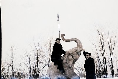 Two men pose atop a large, abstract elephant sculpture in a winter landscape. The man on the elephant’s back wears a peaked c...