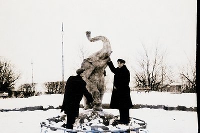 Two men in winter coats inspect a mid-20th-century concrete elephant statue in a snowy park. The sculpture’s dynamic pose and...