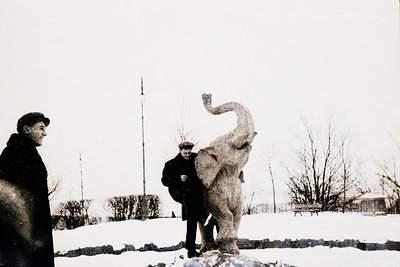 Black-and-white photo of two men posing beside a dynamic elephant sculpture in an urban park. The sculpture’s raised front le...