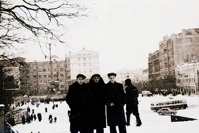 Mid-20th century urban winter scene with three men posing in winter coats and caps. Snow-covered street and ice rink in backg...