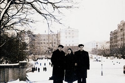 Three individuals pose in winter attire—overcoats, scarves, and hats—on a snow-covered urban square. Soviet-era architecture ...