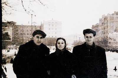 Three individuals pose in winter attire—men in flat caps, woman in a high-collared coat—on a snowy urban street, likely Easte...