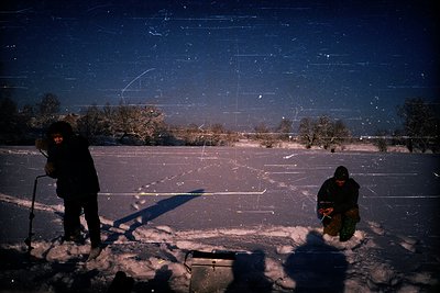 Two figures ice fishing under a starry night sky, silhouetted against a frozen lake. Snow-covered landscape with sparse trees...