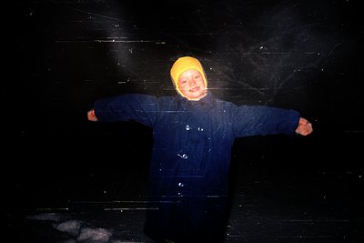 Child in a dark, snowy environment poses with arms outstretched, wearing a bright yellow beanie and dark waterproof coat. Fro...