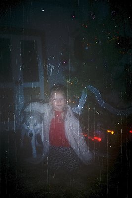 A woman and dog framed by rain-streaked glass, illuminated by indoor light. The woman wears a patterned sweater and holds a d...