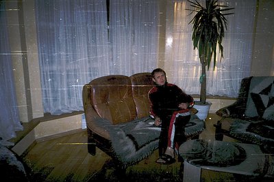 Vintage interior shot featuring a lone individual seated on a worn leather sofa in a dimly lit room. The scene includes a pot...