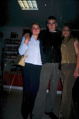 Three individuals pose indoors, likely in a pharmacy or apothecary setting, judging by the shelves of glass bottles behind th...