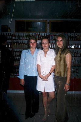 Three women pose indoors in a dimly lit setting, likely a 1970s-80s liquor store. The central figure wears a white short-slee...