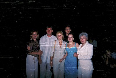 Five adults pose indoors against a dark, textured wall, likely a 1980s–1990s era photo. Men wear dress shirts; women in forma...