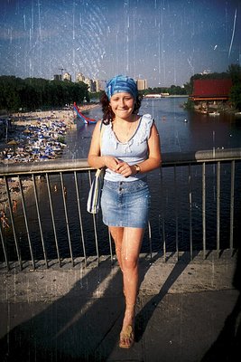A woman in a blue headscarf and denim skirt poses on a wet bridge railing, holding a small bag. Raindrops streak the scene, c...