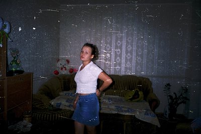 Vintage indoor portrait of a woman in a 1970s-style cropped white top and denim skirt, standing in a dimly lit room with peel...