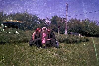 Three individuals in vintage attire sit on grassy hillside, framed by lush greenery and utility poles. Distinctive 1960s fash...
