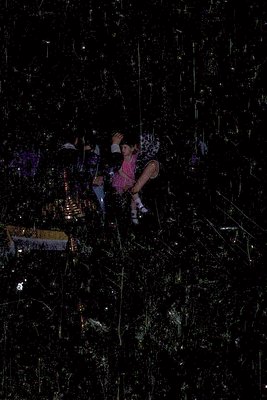A young girl in a pink dress and white socks clings to a suspended net in a dark indoor space, illuminated by scattered light...