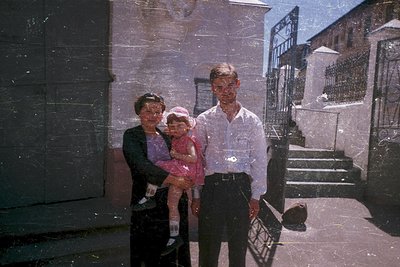 Family portrait in mid-20th century urban setting. Three generations pose on concrete steps beside a wrought-iron gate and br...