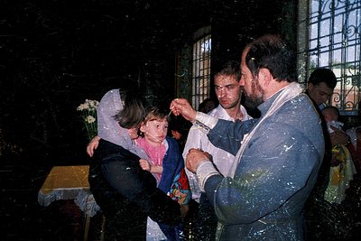 A priest sprinkles holy water on a child during a religious ceremony, likely an Orthodox Christian baptism. The scene capture...