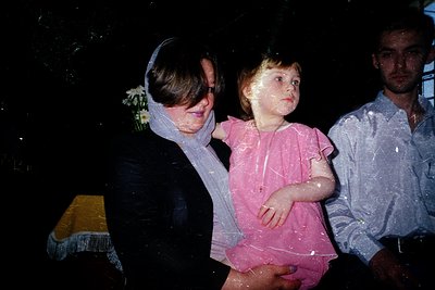 Vintage water splash candid: adult in wet black coat cradles a child in pink dress, both drenched. Man in light shirt stands ...