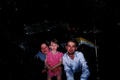 Vintage black-and-white photo of three individuals standing under a decorative umbrella in rain, likely 1960s–1970s. Woman in...