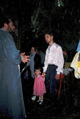 A priest in liturgical vestments blesses a young girl in a pink dress during an outdoor evening ceremony. Adults in casual 19...