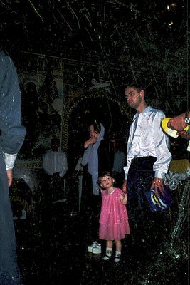 A dimly lit indoor scene featuring three figures in a traditional or folk setting. A man in a white shirt and tie holds a chi...