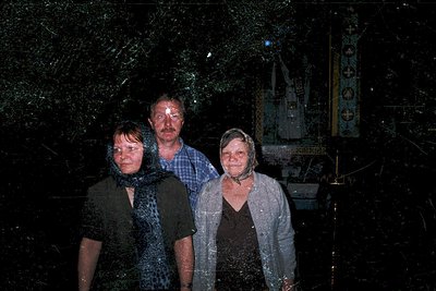 Three individuals pose under heavy rain in a rural setting, likely Eastern Europe, 1970s-1980s. The man wears a checkered shi...
