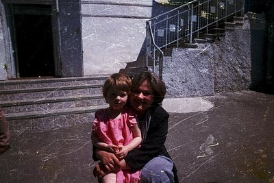 Vintage candid of adult and child seated on stone steps, likely 1970s–1980s. The adult wears a dark jacket and blue jeans; th...
