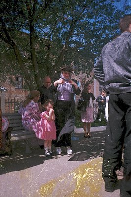 Mid-20th century family moment in a park—man in suit claps hands, children laugh as water splashes from a fountain. Brick bui...