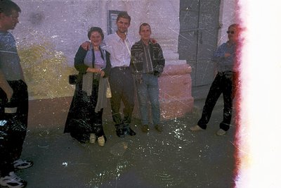 Vintage group photo in urban floodwaters, 1970s-80s. Four individuals pose in wet clothing—men in jackets, jeans, and boots; ...