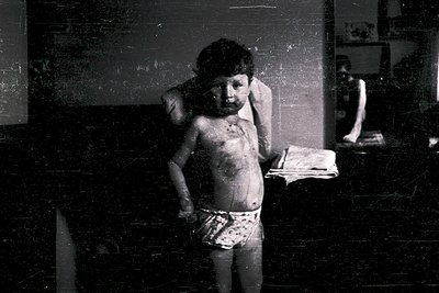 Young boy in diaper stands in dimly lit room, holding crumpled paper. Peeling wallpaper and aged wall texture suggest mid-20t...
