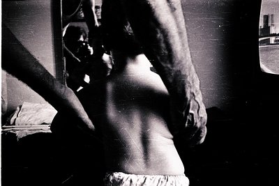 Black-and-white candid shot of a person in a mirrored room, captured from behind. Reflections reveal a partial view of a bed ...