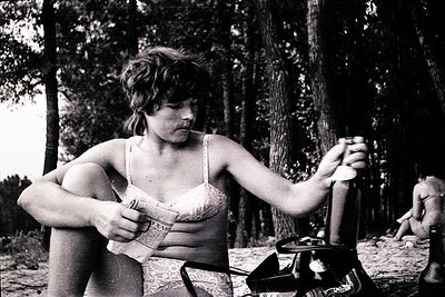 Vintage black-and-white photo of a woman in a bikini top holding a rolled-up garment, standing beside a portable stove on a s...