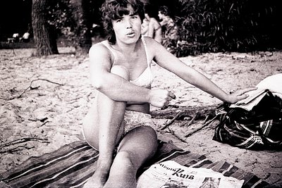 Black-and-white beach scene featuring a seated woman in a one-piece swimsuit, posed on a striped towel. A newspaper with Cyri...