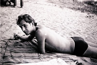 Mid-century beach scene: shirtless man in swim trunks rests on a striped towel on sandy shore, 1960s-70s. Classic retro beach...