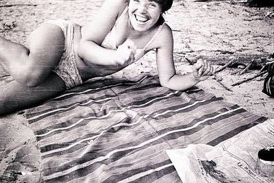 Mid-20th century beach scene: Woman in vintage swimsuit lounges on striped beach towel, smiling at camera. Sandy, natural set...