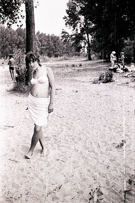 Mid-century beach scene featuring a woman in a minimalist, knee-length sarong. She stands barefoot on sandy terrain, surround...