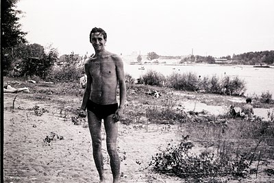 Mid-century beachgoer in minimalist swim trunks poses by a lakeside, likely 1950s–1960s. Overgrown vegetation and distant boa...