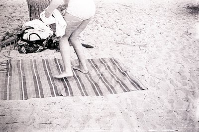 Mid-century beach scene: barefoot child in shorts steps onto a striped woven mat on sandy shore. Packed bag and towel beside ...