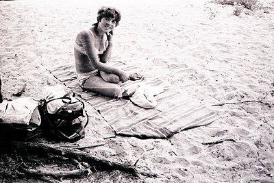Mid-century beach scene featuring a woman seated on a striped towel, holding a sun hat. Beside her lies a vintage backpack an...