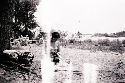 Black-and-white mid-20th century camp scene by a waterfront. A woman in a sleeveless dress and headscarf tends a small fire o...