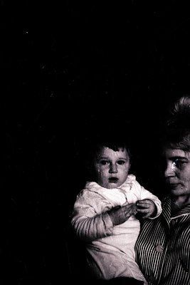 Black-and-white portrait of an adult holding a young child indoors, likely mid-20th century. The adult wears a striped shirt,...