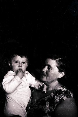 Vintage black-and-white photo of an elderly woman holding an infant, both indoors. The woman wears a patterned blouse with a ...