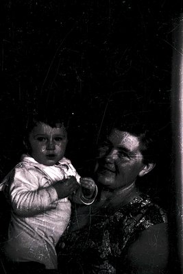 Vintage black-and-white portrait of an elderly woman holding a young child indoors, likely mid-20th century. The woman’s patt...