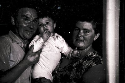 Black-and-white candid of elderly couple holding a baby indoors, likely mid-20th century. Man in light shirt, woman in patter...
