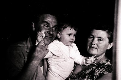 Black-and-white portrait of an elderly man and woman holding a baby indoors, likely mid-20th century. The man’s hand gestures...