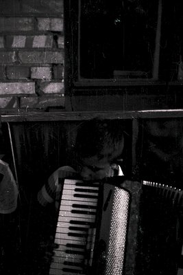 Close-up of a person playing an accordion in dim lighting, framed by a rustic brick wall. The vintage monochrome tone suggest...