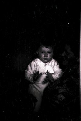 Vintage sepia-toned portrait of a young child in a long-sleeve onesie, seated indoors near a curtained window. The child’s ex...