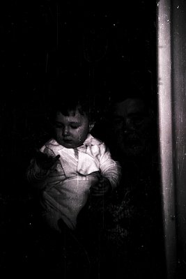 Vintage black-and-white photo of a young child pressing their face against a windowpane, hands gripping the sill. Soft indoor...
