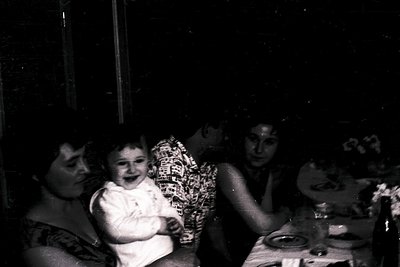 Vintage black-and-white photo of a family moment: a woman cradles a laughing toddler, another adult smiles beside them at a t...
