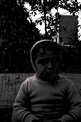 Black-and-white portrait of a man in a knit sweater with a geometric, abstract mask overlay. Urban setting with blurred folia...