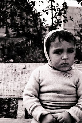 Vintage black-and-white portrait of a young child in a striped sweater and knitted cap, seated on a weathered wooden bench. T...